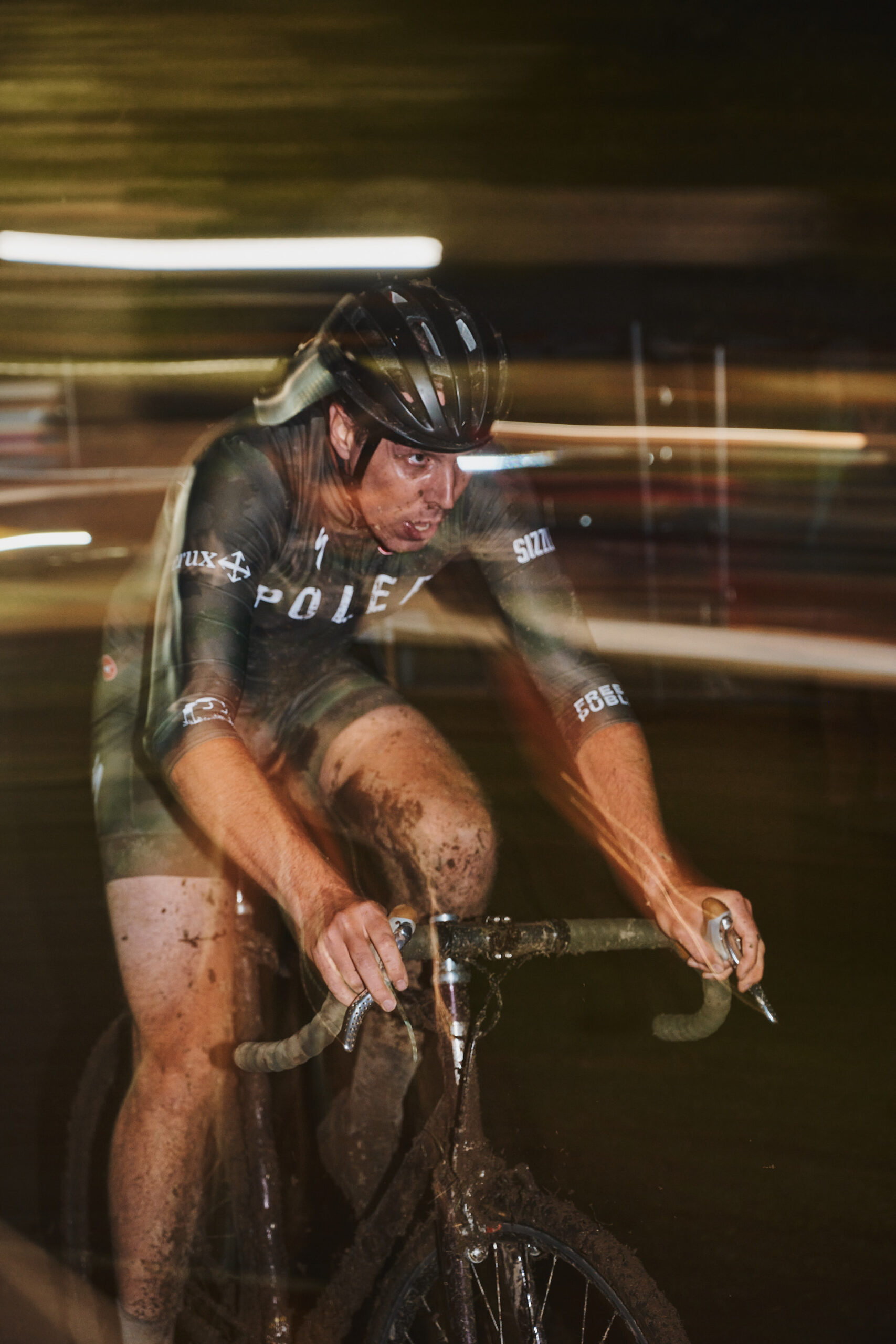 A Cyclocross Racer speeds by at night with light trails from a long exposure loom in the background at Portland Trophy Cup 2023