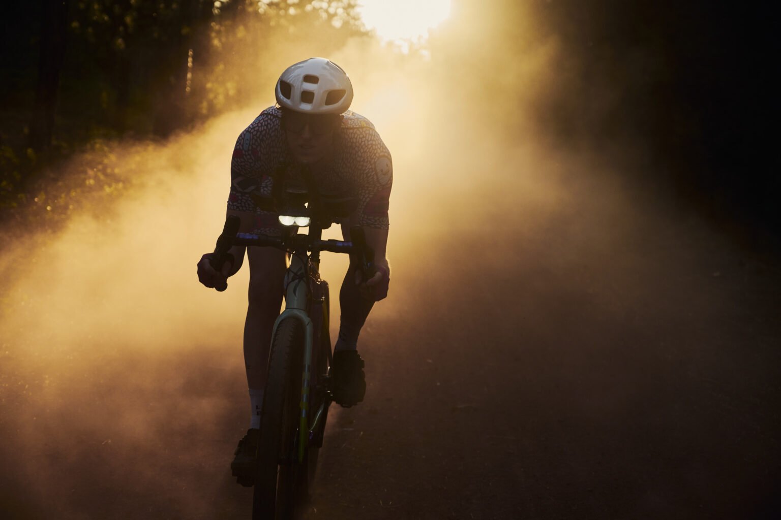 A gravel cyclist emerges from a backlit dust cloud as a silhouette.